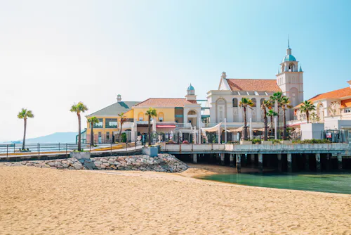 A sunny beach with golden sand leads to a pier with elegant, Mediterranean-style buildings, palm trees, and a clear blue sky in the background.