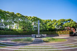 A circular memorial plaza with a tall stone obelisk at its center, surrounded by green trees and blue sky, with steps leading up to the monument and curved walkways encircling the area.