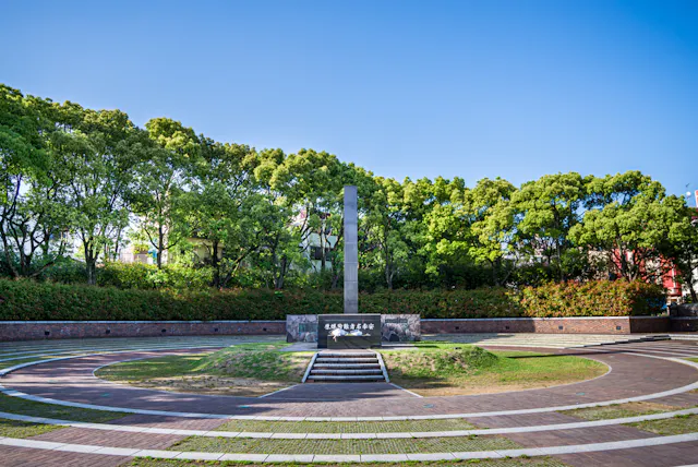 A circular memorial plaza with a tall stone obelisk at its center, surrounded by green trees and blue sky, with steps leading up to the monument and curved walkways encircling the area.