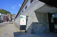 A stone building with a sign reading “Nagasaki Museum of History and Culture” stands beside a quiet street under a blue sky. A few people walk along the sidewalk near the entrance.