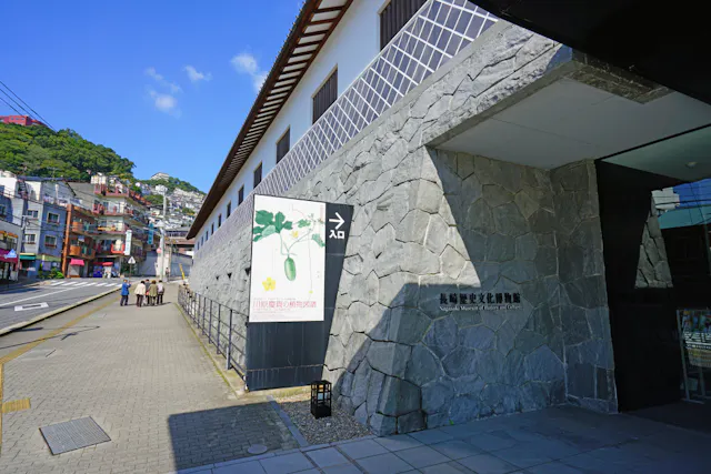A stone building with a sign reading “Nagasaki Museum of History and Culture” stands beside a quiet street under a blue sky. A few people walk along the sidewalk near the entrance.