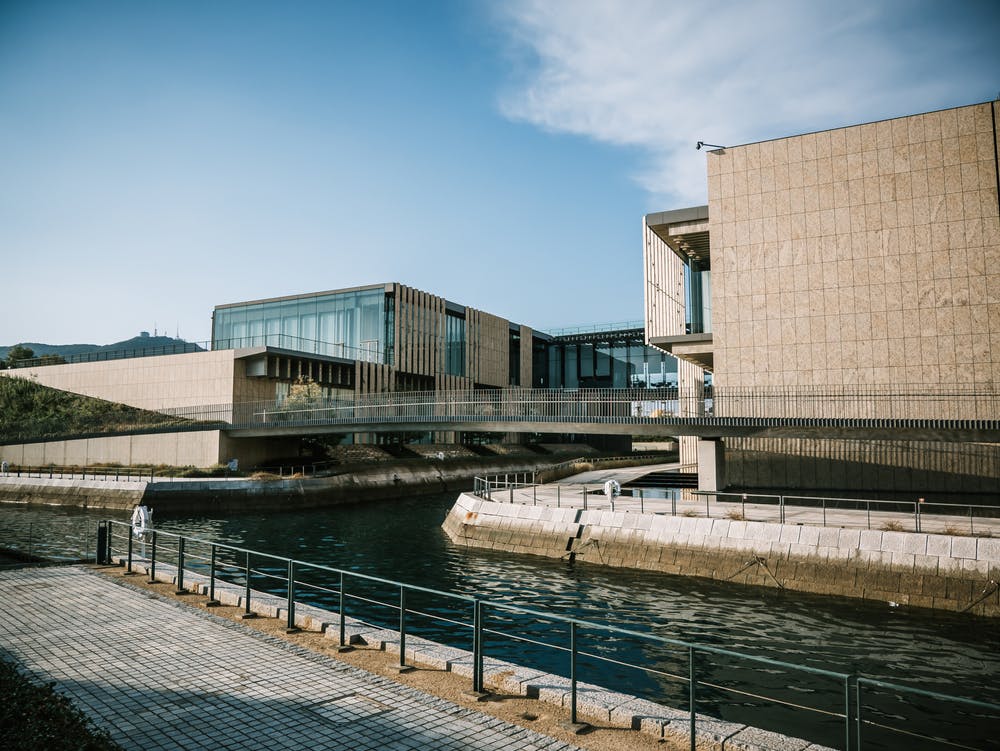 Modern buildings with large glass windows and beige stone facades sit beside a calm canal, connected by elevated walkways. A paved path with railings runs along the water under a clear blue sky.