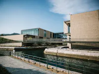Modern buildings with large glass windows and beige stone facades sit beside a calm canal, connected by elevated walkways. A paved path with railings runs along the water under a clear blue sky.