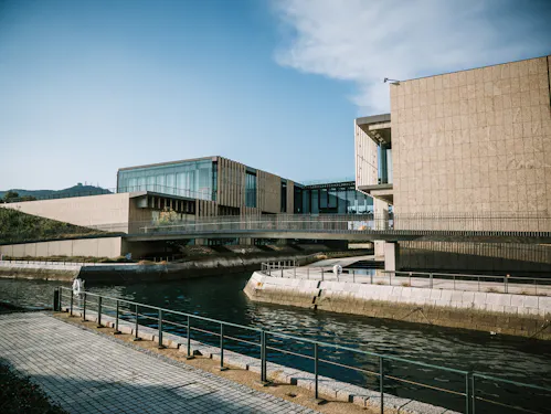Modern buildings with large glass windows and beige stone facades sit beside a calm canal, connected by elevated walkways. A paved path with railings runs along the water under a clear blue sky.
