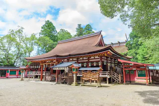 A traditional Japanese Shinto shrine with a curved roof, wooden structure, and decorative details, surrounded by trees and a bright sky. Wooden ema plaques hang near the entrance.