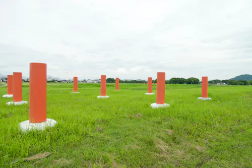 Several tall, orange cylindrical posts with white bases are evenly spaced across a grassy field under a cloudy sky, with distant trees and hills in the background.