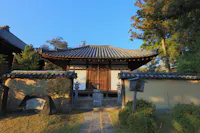 A traditional Japanese building with a tiled roof stands behind a stone pathway, low beige walls, lanterns, and lush greenery, all bathed in warm sunlight.