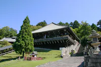 A traditional Japanese wooden temple stands elevated on a hill, surrounded by green trees and grass, with a stone staircase and lanterns leading up to the entrance under a clear blue sky.