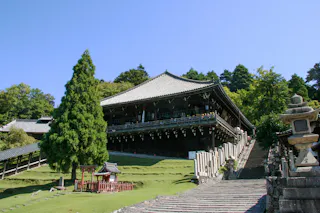 A traditional Japanese wooden temple stands elevated on a hill, surrounded by green trees and grass, with a stone staircase and lanterns leading up to the entrance under a clear blue sky.