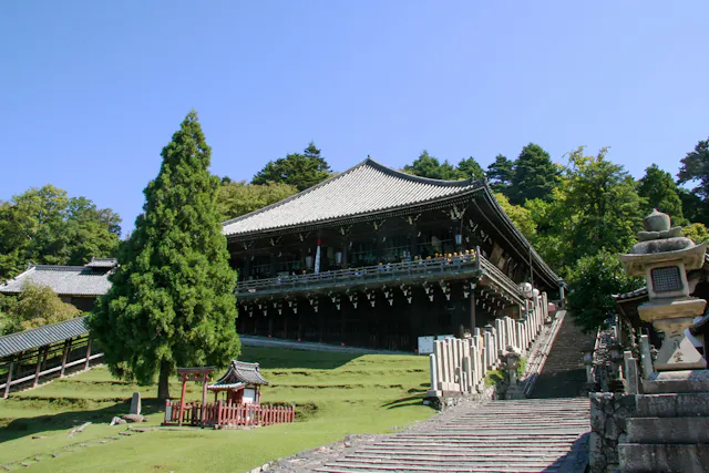 A traditional Japanese wooden temple stands elevated on a hill, surrounded by green trees and grass, with a stone staircase and lanterns leading up to the entrance under a clear blue sky.