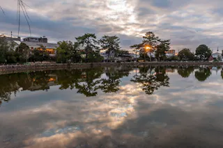 A calm lake reflects trees, buildings, and a cloudy sky at sunset, with the sun partially visible through the trees near the water’s edge.