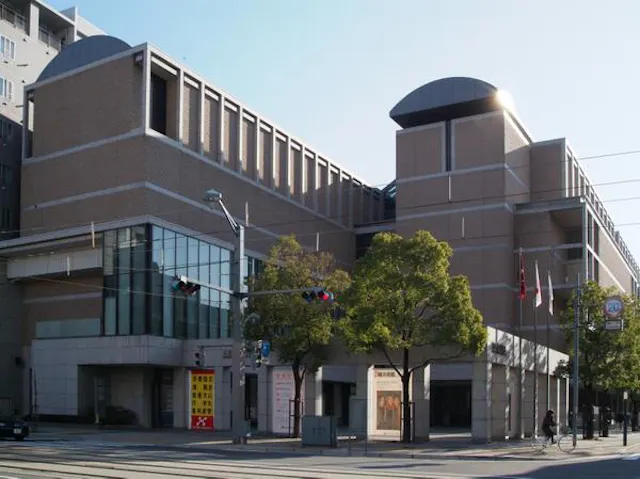 A modern, multi-story brick building with large windows and geometric shapes. Trees line the sidewalk, and two people walk nearby. Traffic lights and street signs are visible in the foreground.