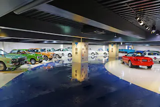 A showroom displaying a lineup of classic cars in various colors including green, orange, white, blue, and red, all parked in a sleek, modern indoor space with glossy floors and spotlights above.