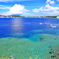 Cape Maeda (Maeda Flats) A vibrant coastal scene with clear blue water, boats floating offshore, and many people swimming and snorkeling near the reef. Lush greenery and distant buildings are visible under a bright, partly cloudy sky.