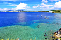 A vibrant coastal scene with clear blue water, boats floating offshore, and many people swimming and snorkeling near the reef. Lush greenery and distant buildings are visible under a bright, partly cloudy sky.
