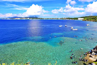 A vibrant coastal scene with clear blue water, boats floating offshore, and many people swimming and snorkeling near the reef. Lush greenery and distant buildings are visible under a bright, partly cloudy sky.