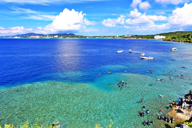 A vibrant coastal scene with clear blue water, boats floating offshore, and many people swimming and snorkeling near the reef. Lush greenery and distant buildings are visible under a bright, partly cloudy sky.