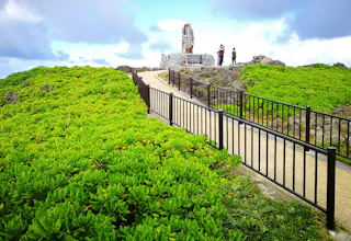 A paved walkway with black railings winds through lush green bushes, leading to a stone monument at the top of a small hill. Three people stand near the monument under a partly cloudy sky.