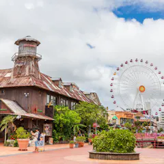 Mihama American Village A rustic, weathered building surrounded by plants stands in the foreground, with a large Ferris wheel and city buildings visible in the background under a partly cloudy sky.