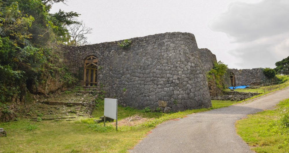 A stone wall and arched gateways form part of the ruins of an ancient castle, surrounded by greenery and a paved path, under a partly cloudy sky.