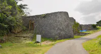 A stone wall and arched gateways form part of the ruins of an ancient castle, surrounded by greenery and a paved path, under a partly cloudy sky.