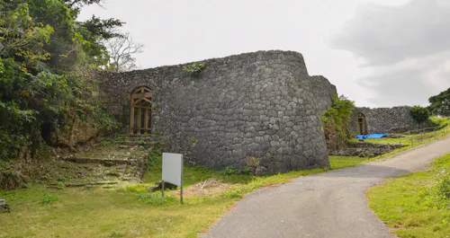 A stone wall and arched gateways form part of the ruins of an ancient castle, surrounded by greenery and a paved path, under a partly cloudy sky.