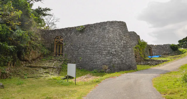 A stone wall and arched gateways form part of the ruins of an ancient castle, surrounded by greenery and a paved path, under a partly cloudy sky.
