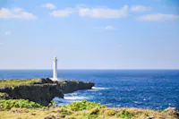 A white lighthouse stands on a rocky cliff overlooking a deep blue ocean under a clear sky with a few clouds. Green vegetation grows on the cliff and in the foreground.
