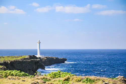 A white lighthouse stands on a rocky cliff overlooking a deep blue ocean under a clear sky with a few clouds. Green vegetation grows on the cliff and in the foreground.