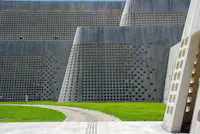 Modern building with tall concrete walls featuring a grid of square openings, set beside a curved pathway and a green lawn under natural daylight.