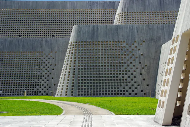 Modern building with tall concrete walls featuring a grid of square openings, set beside a curved pathway and a green lawn under natural daylight.