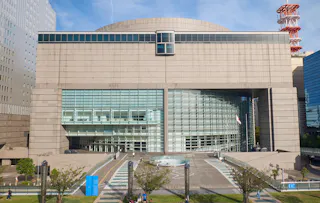 A large modern building with a curved glass facade and beige concrete exterior, featuring an elevated central walkway, trees, and people walking in the surrounding plaza.