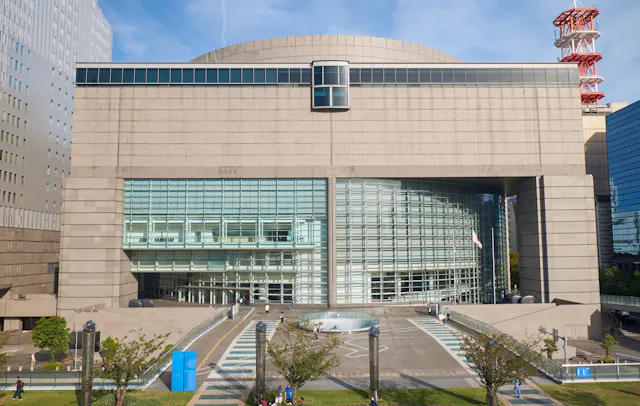 A large modern building with a curved glass facade and beige concrete exterior, featuring an elevated central walkway, trees, and people walking in the surrounding plaza.