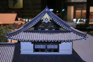 A detailed model of a traditional Japanese building roof, featuring intricate tile work and ornate gold decorations under the gable, with a barred window below the roof section.