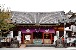 A traditional Japanese shrine with a tiled roof, purple banners, lanterns, and stone statues at the entrance. A tree stands to the left, and various signs and notices are displayed around the building.