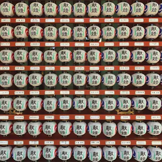 Rows of traditional Japanese sake barrels, each decorated with calligraphy and arranged neatly in a wooden display. White labels with Japanese writing are placed below each barrel.