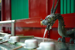 A bronze dragon fountain spouts water into a basin, with metal ladles resting on the edge. The background features red and green vertical panels, giving a traditional Japanese aesthetic.