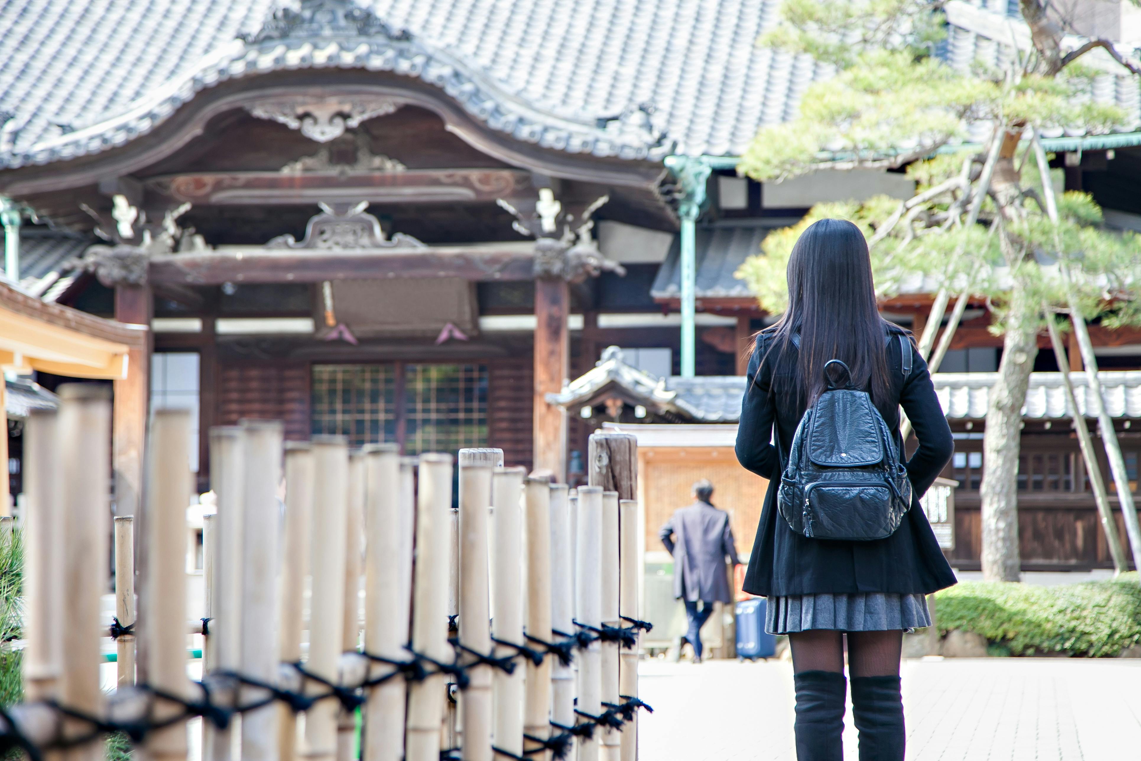 A person with long hair, wearing a black outfit and backpack, stands facing a traditional Japanese temple. Bamboo railings and another person are visible in the background.
