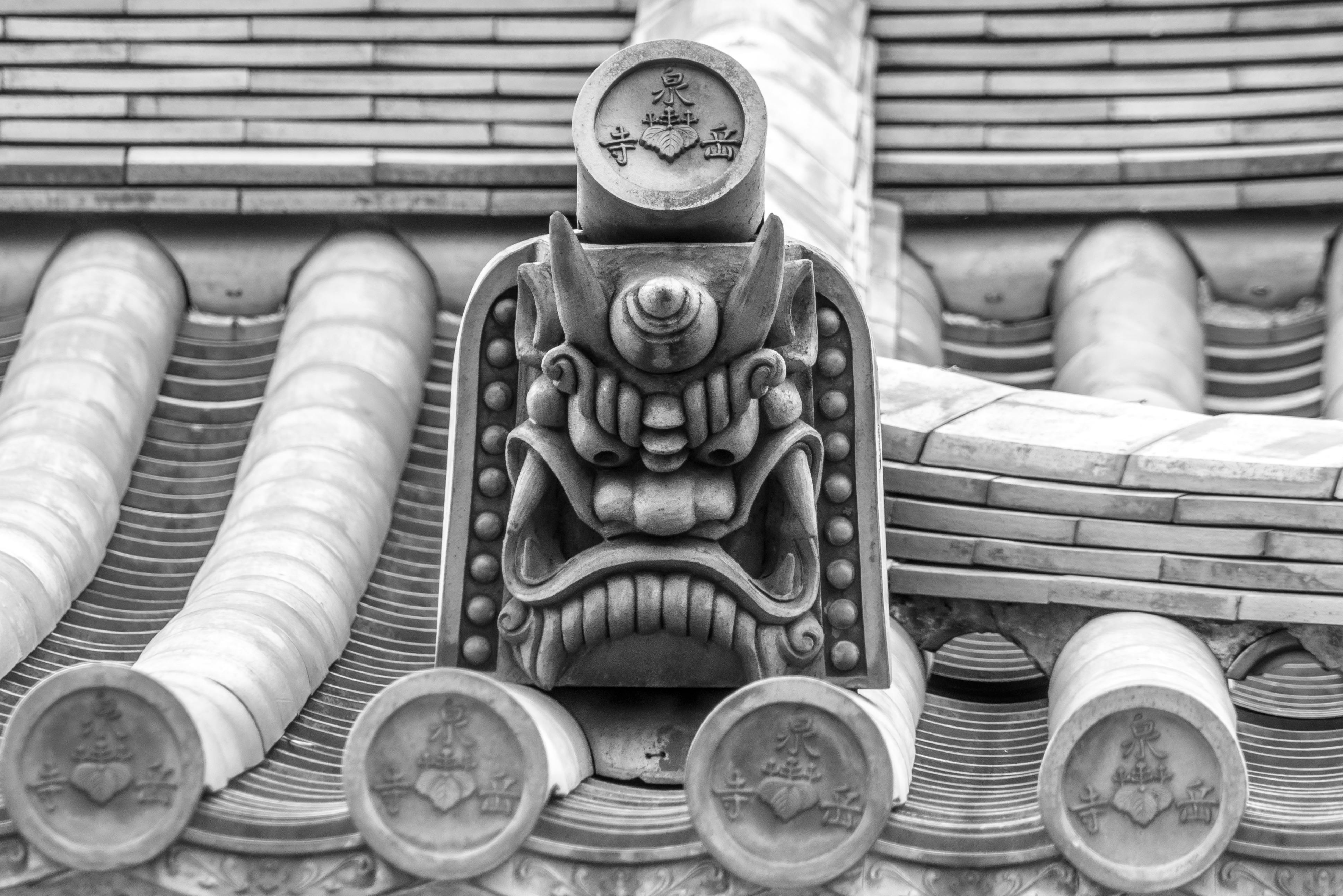 Close-up of an ornate, fierce-looking roof tile ornament on a traditional Japanese building, featuring a detailed mythical face with sharp eyes and teeth. The image is in black and white.