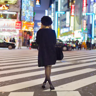 A person in a black outfit and heels walks across a striped crosswalk at night, surrounded by bright city lights and neon signs in an urban area.