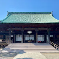 Gokokuji Temple A traditional Japanese temple with a green tiled roof, wide steps leading up to the entrance, wooden pillars, and lanterns, set against a clear blue sky.