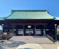 A traditional Japanese temple with a green tiled roof, wide steps leading up to the entrance, wooden pillars, and lanterns, set against a clear blue sky.