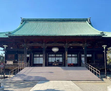 A traditional Japanese temple with a green tiled roof, wide steps leading up to the entrance, wooden pillars, and lanterns, set against a clear blue sky.