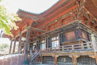 A traditional Japanese wooden temple with intricate carvings and a large overhanging roof, surrounded by greenery and steps leading up to the entrance.