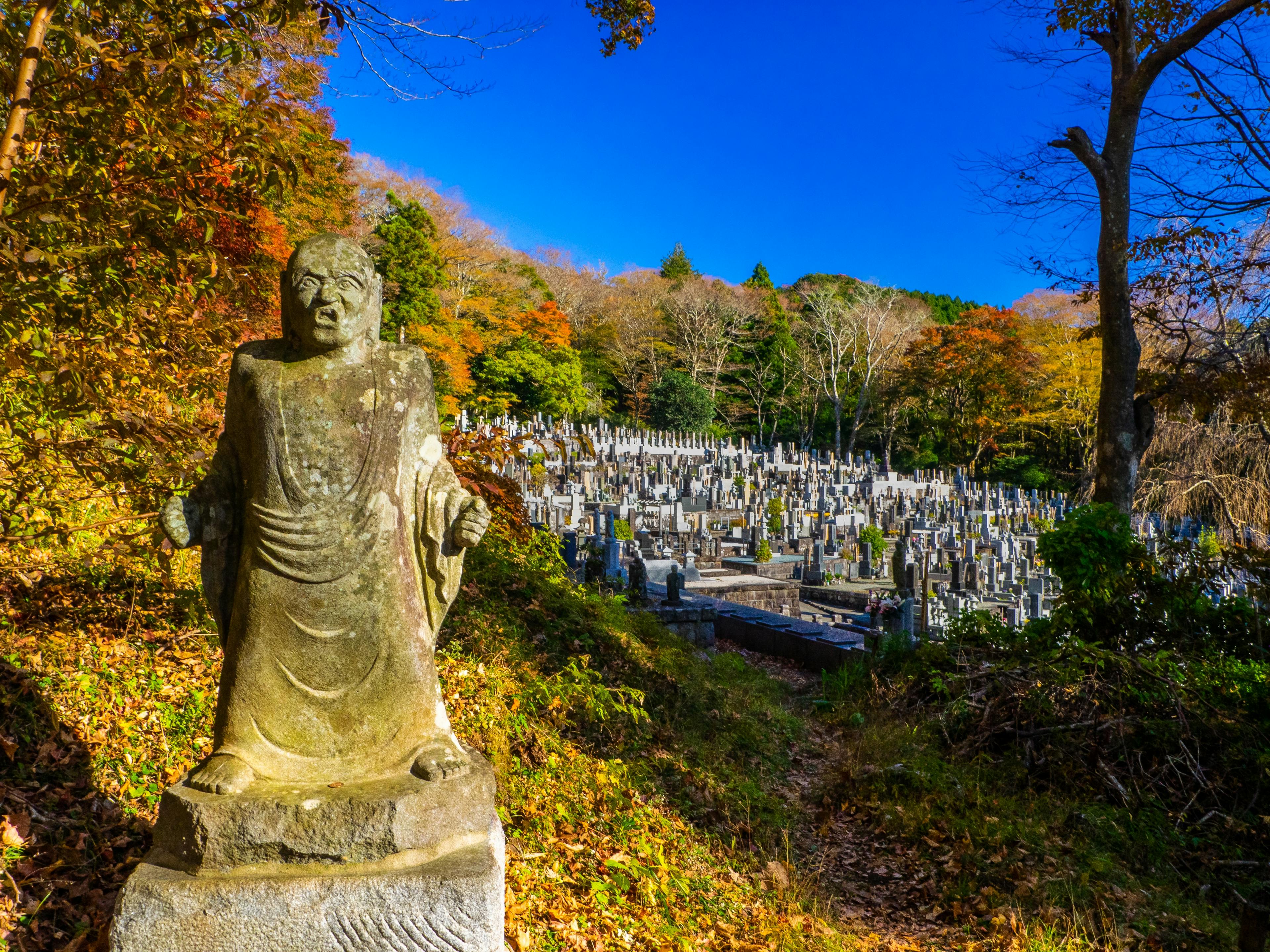 A stone statue stands near a path overlooking a hillside Japanese cemetery with many gravestones, surrounded by autumn trees under a clear blue sky.