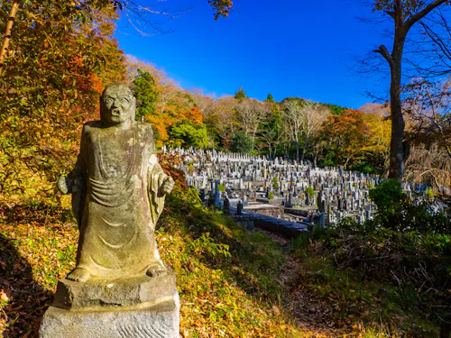 Choanji A stone statue stands near a path overlooking a hillside Japanese cemetery with many gravestones, surrounded by autumn trees under a clear blue sky.