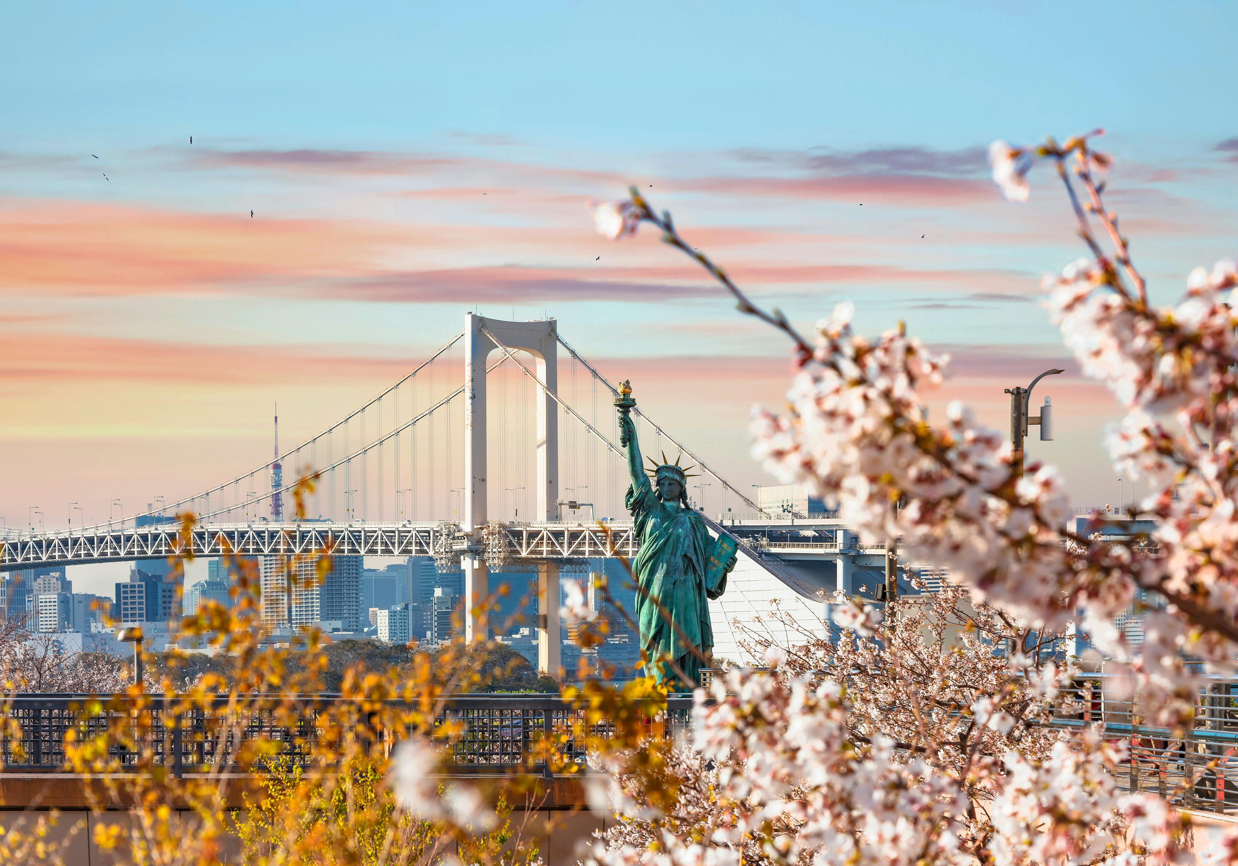 Cherry blossoms in the foreground frame a replica of the Statue of Liberty, with a large suspension bridge and a city skyline visible in the background under a colorful sunset sky.