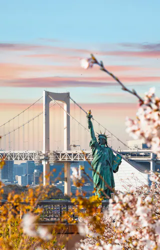 Odaiba Seaside Park Cherry blossoms in the foreground frame a replica of the Statue of Liberty, with a large suspension bridge and a city skyline visible in the background under a colorful sunset sky.