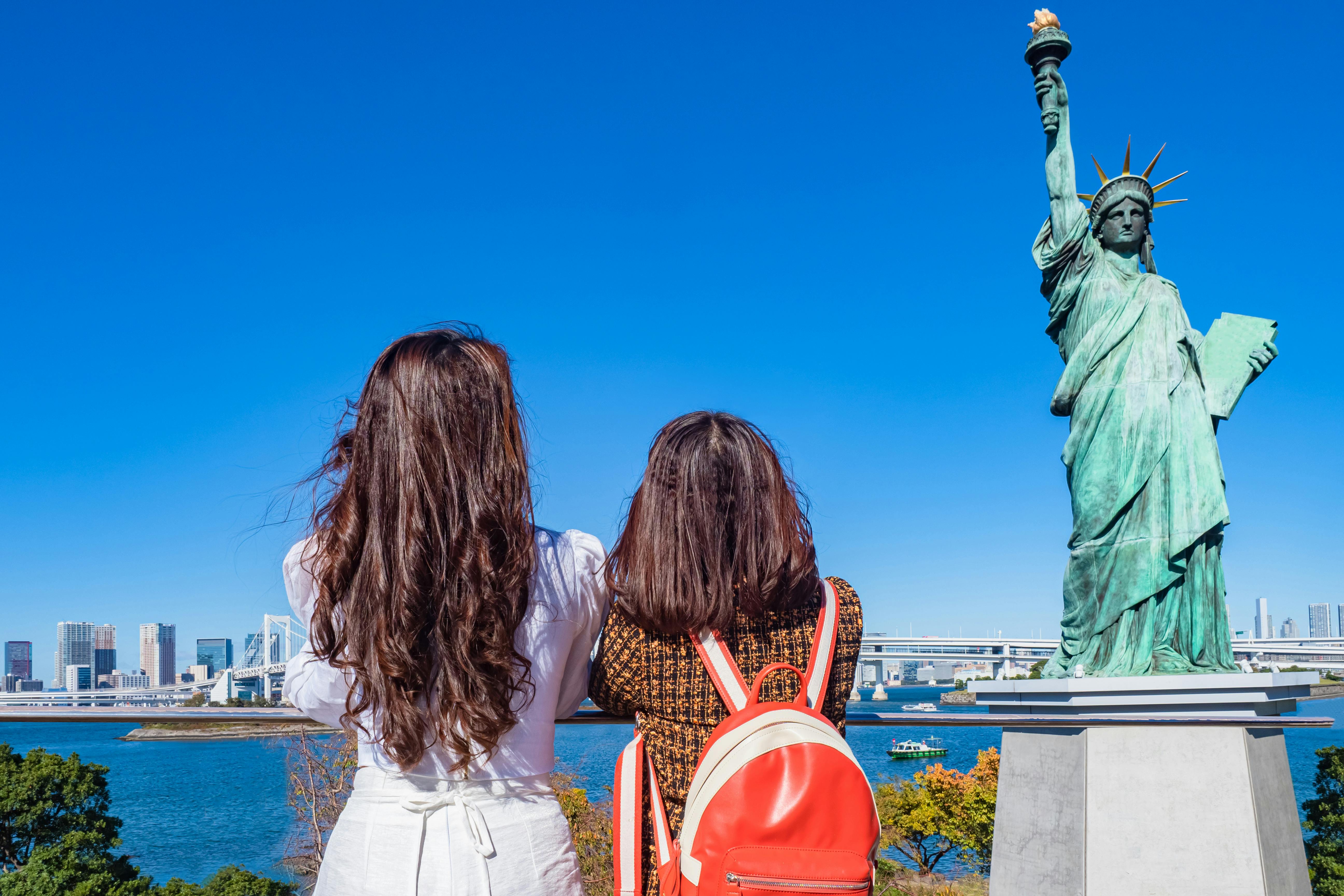 Two women with long hair, seen from behind, stand near a waterfront looking at a replica of the Statue of Liberty. City buildings and a bridge are visible in the background under a clear blue sky.