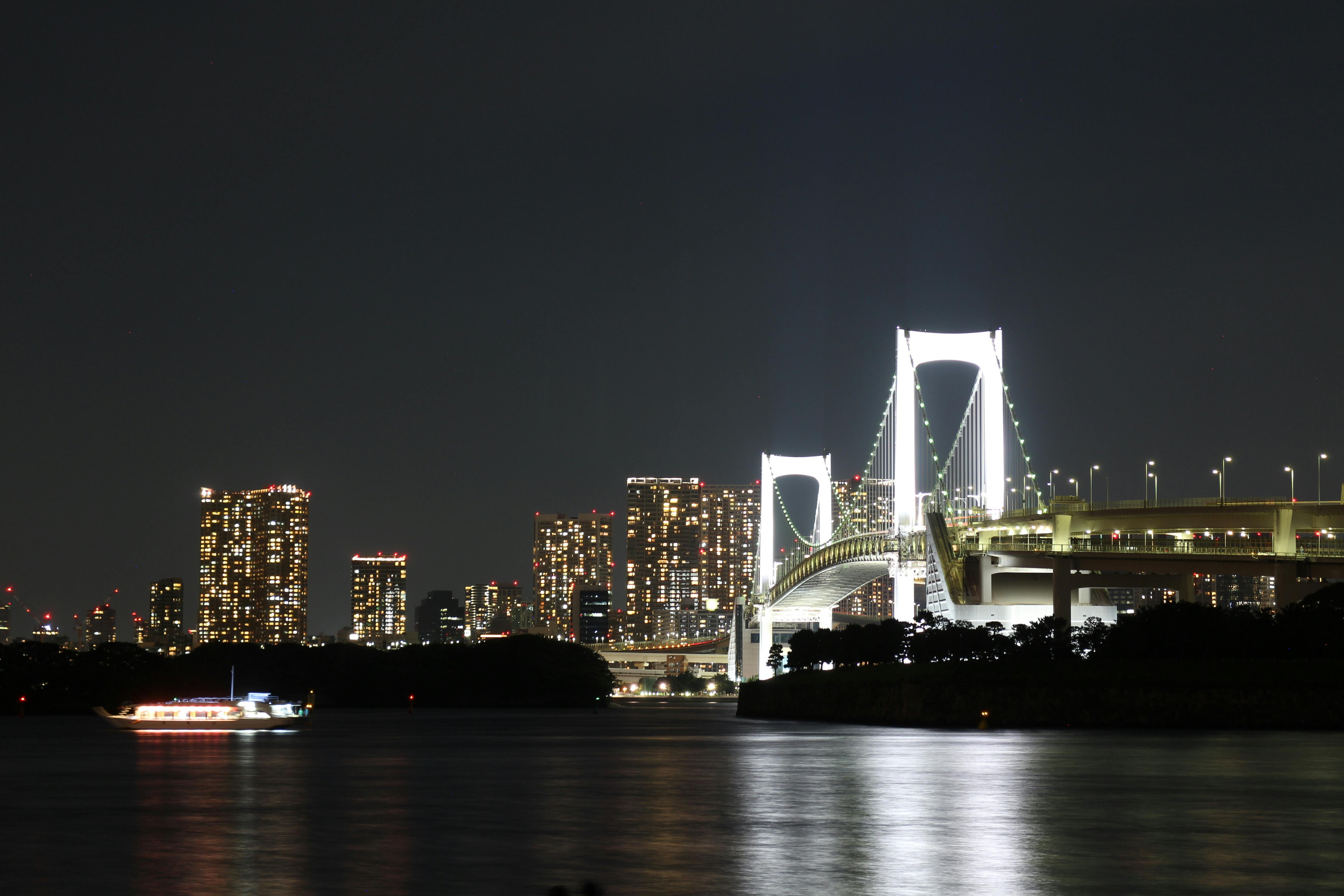 A brightly lit suspension bridge stretches over a calm river at night, with a boat illuminated on the water and tall city buildings glowing in the background.